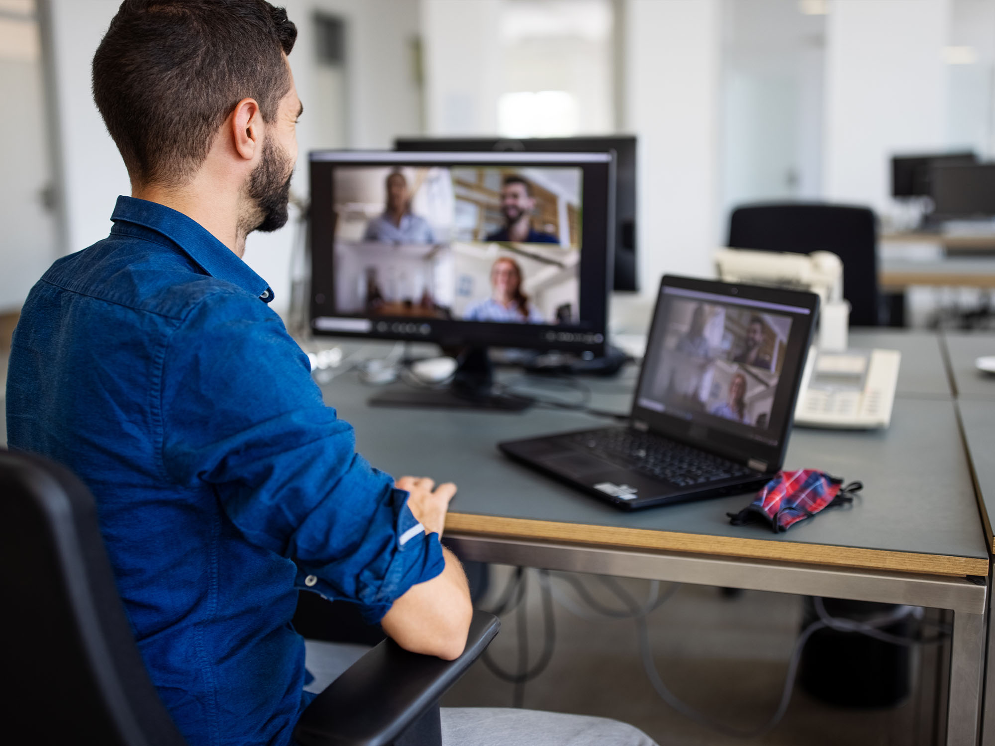 A man sits in front of his computer monitor and laptop, each showing a Zoom meeting room with 4 participants