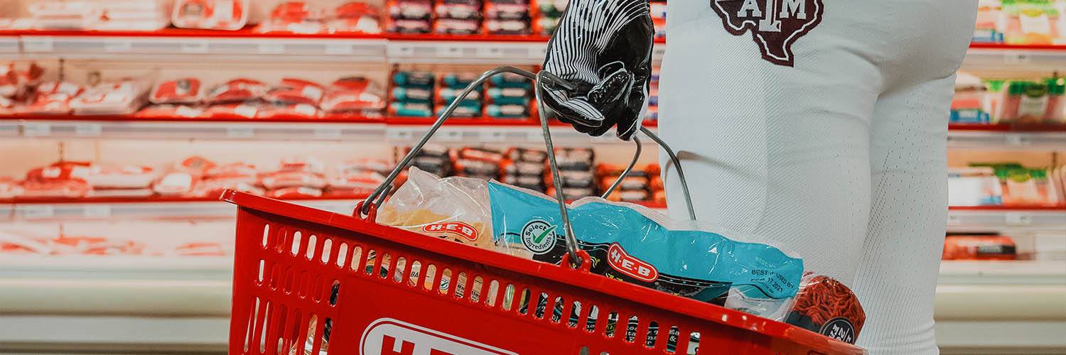 a Texas A&M yell leader holds a basket of groceries while standing in front of the meat section at HEB