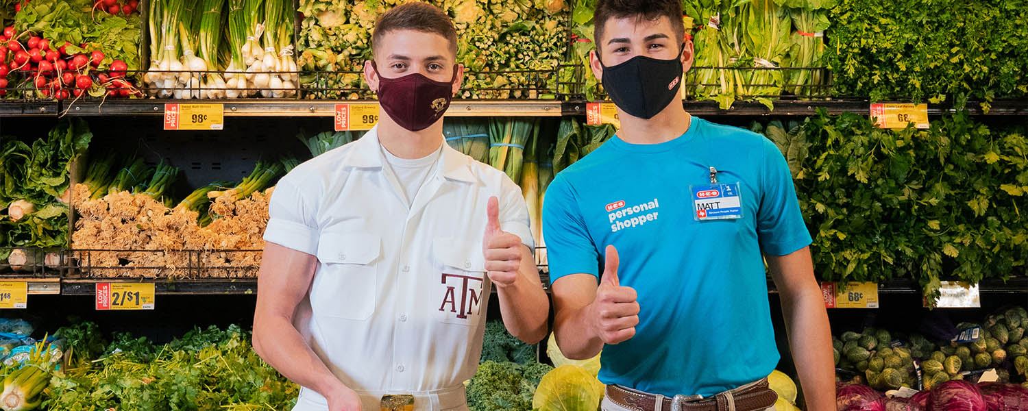 A Texas A&M yell leader holding a basket of groceries stands next to an HEB employee