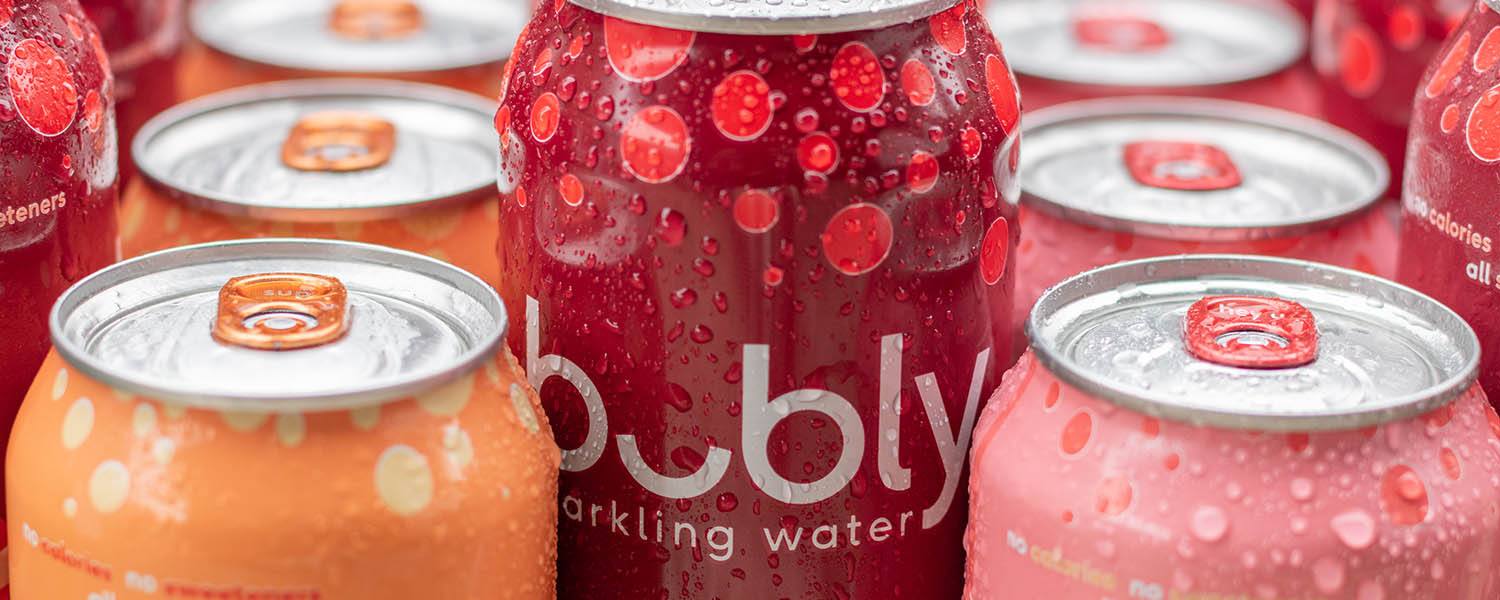 A red can of Bubly is standing above rows of orange and pink Bubly cans.