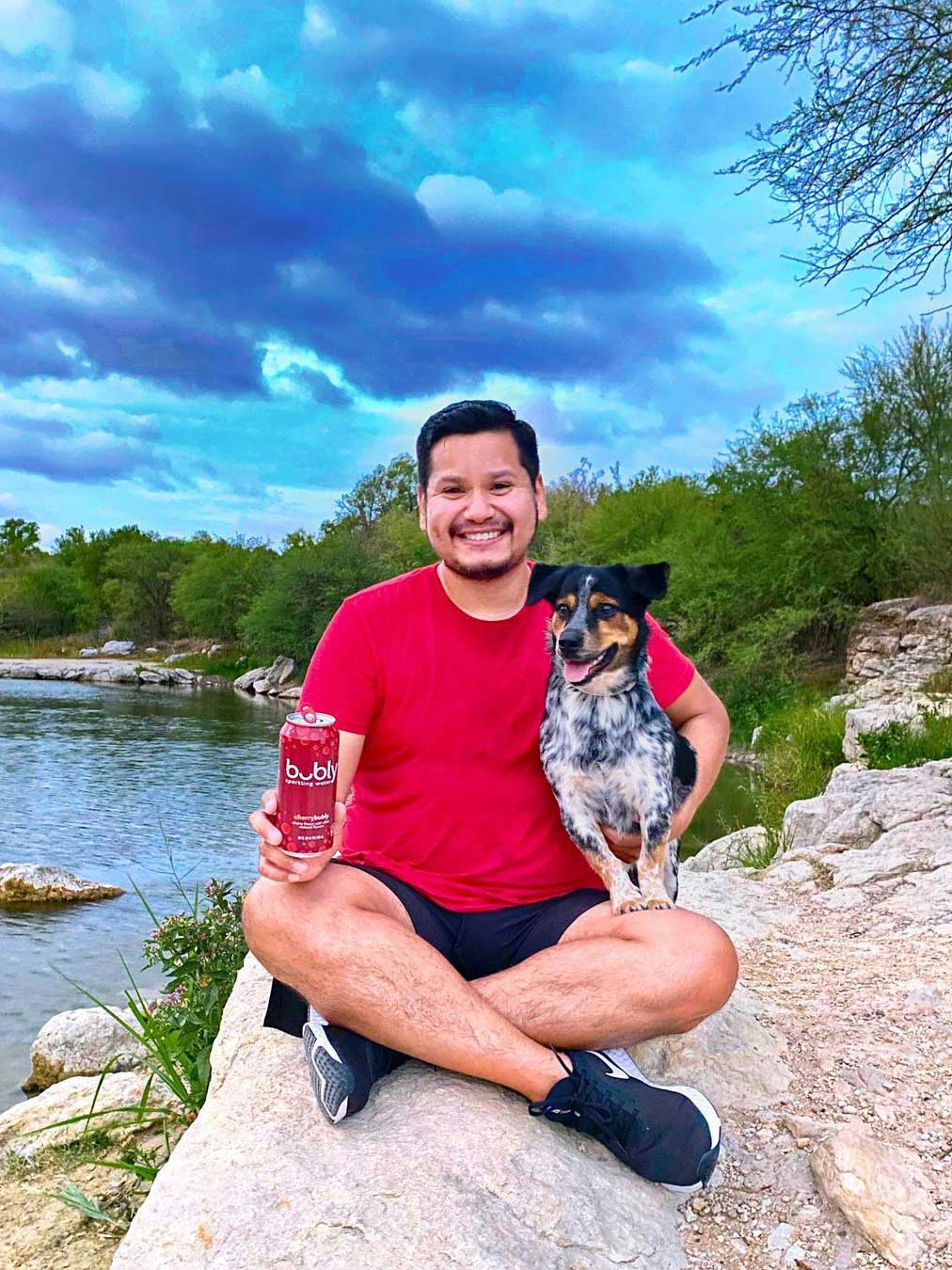 A man sits with his dog on a rock while holding a can of bubly; behind him is a lake, trees, and clouds