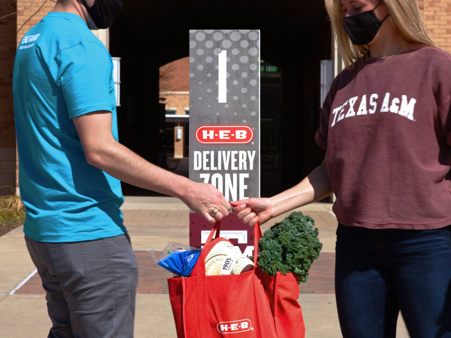 H-E-B employee hands a Texas A&M student their grocieries in front of a Delivery Zone sign on campus